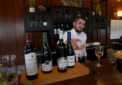 A bartender pours wine at a bar with five bottles displayed on the counter, set against a warm-toned background with shelving.