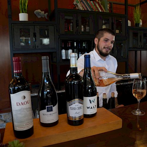 A bartender pours wine at a bar with five bottles displayed on the counter, set against a warm-toned background with shelving.