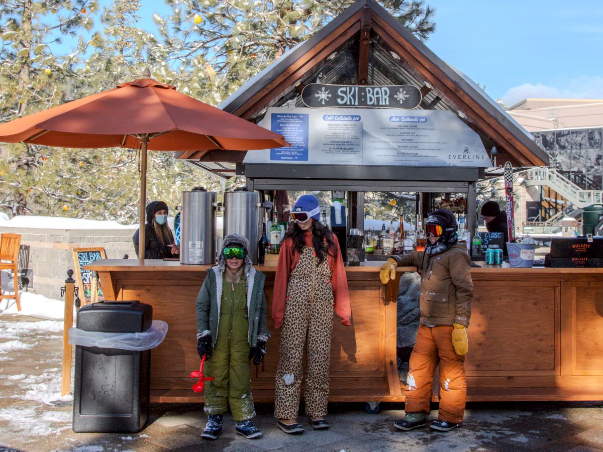 A ski bar with people in winter clothing standing in front, surrounded by snow and pine trees, under a blue sky.