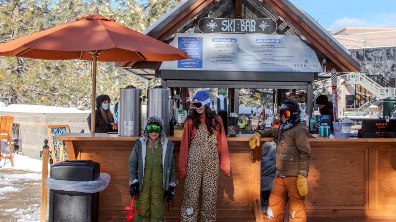 A ski bar with people in winter clothing standing in front, surrounded by snow and pine trees, under a blue sky.