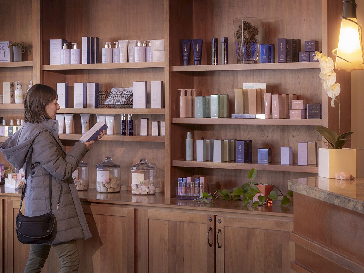 A woman browses a shelf of skincare/perfume bottles in a cozy store, holding a boxed product.