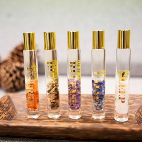 Five gold-capped perfume/serum bottles with colored gemstone-like pieces, arranged on a wooden tray.