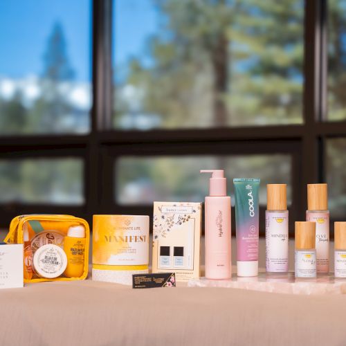 A display of skincare and body-care products&mdash;bottles, lotions, and candles&mdash;on a table by a window with a blurred outdoor view.