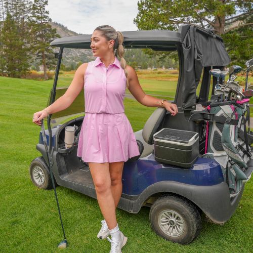 A woman in a pink outfit stands beside a golf cart loaded with clubs on a sunny green course.