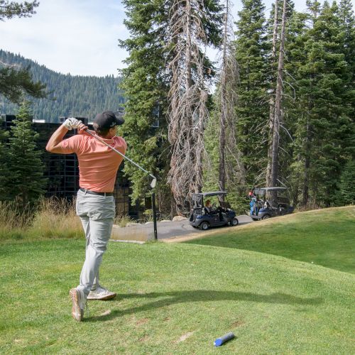 A golfer swings a club on a lush green course, with golf carts parked nearby and tall pines and mountains in the background.