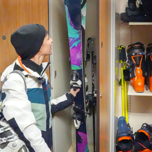 A skier in a locker checks skis while orange ski boots and gear hang on the wall nearby.