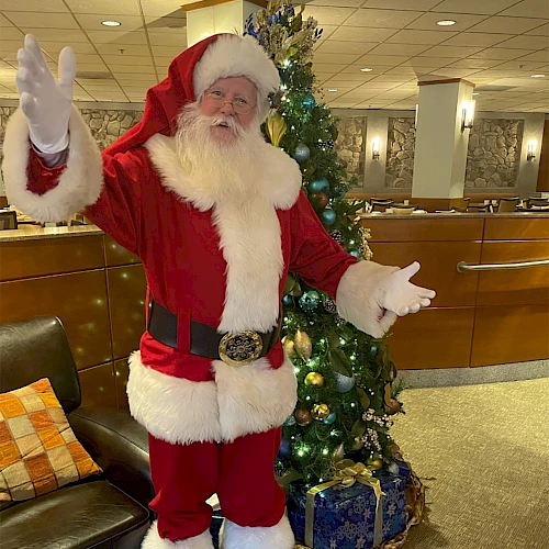 Santa Claus in a red suit with white trim, standing by a decorated Christmas tree in an indoor lobby, waving hello to the camera.