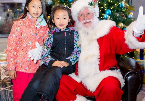 Two children pose with Santa Claus in front of a festive Christmas tree, dressed warmly in winter clothes, all smiling for the photo.