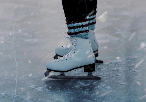 A person wearing white ice skates and blue-striped socks is gliding on an ice rink, with visible ice markings and a snowy texture.