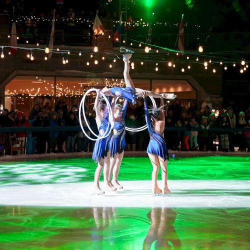 A group of ice skaters performs a routine under green lights, with one skater lifted while holding hoops, surrounded by an audience.