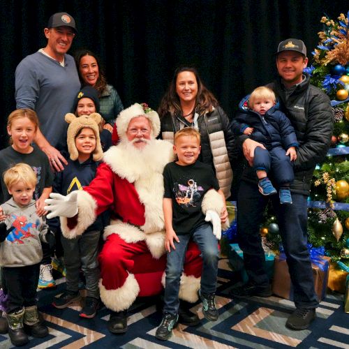 A group of people, including children, are posing with Santa Claus next to a decorated Christmas tree. Everyone looks happy and festive.