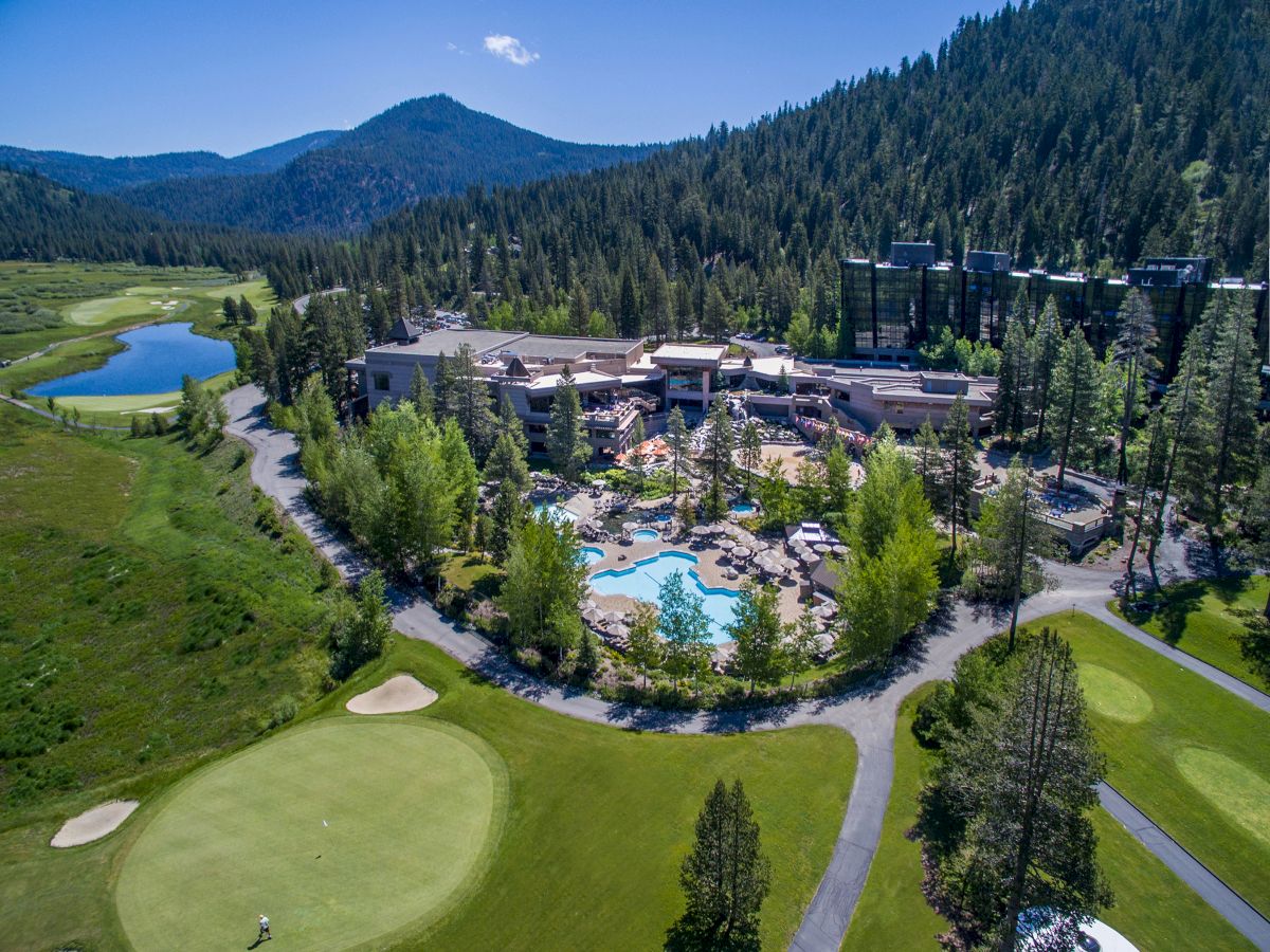 An aerial view of a resort with a central pool area, surrounded by trees, golf courses, and mountains in the background.