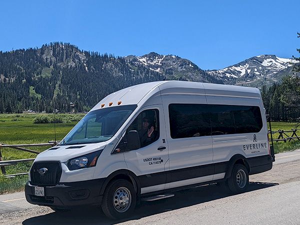 A white shuttle van parked on a mountain road beside green fields, with snowy peaks and trees in the background.