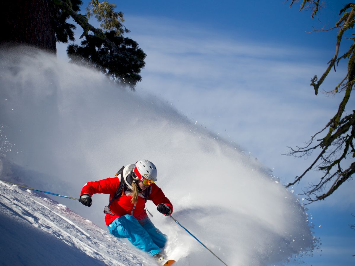 A skier in a red jacket and blue pants makes a sharp turn on a snowy slope, creating a spray of snow under a clear blue sky.