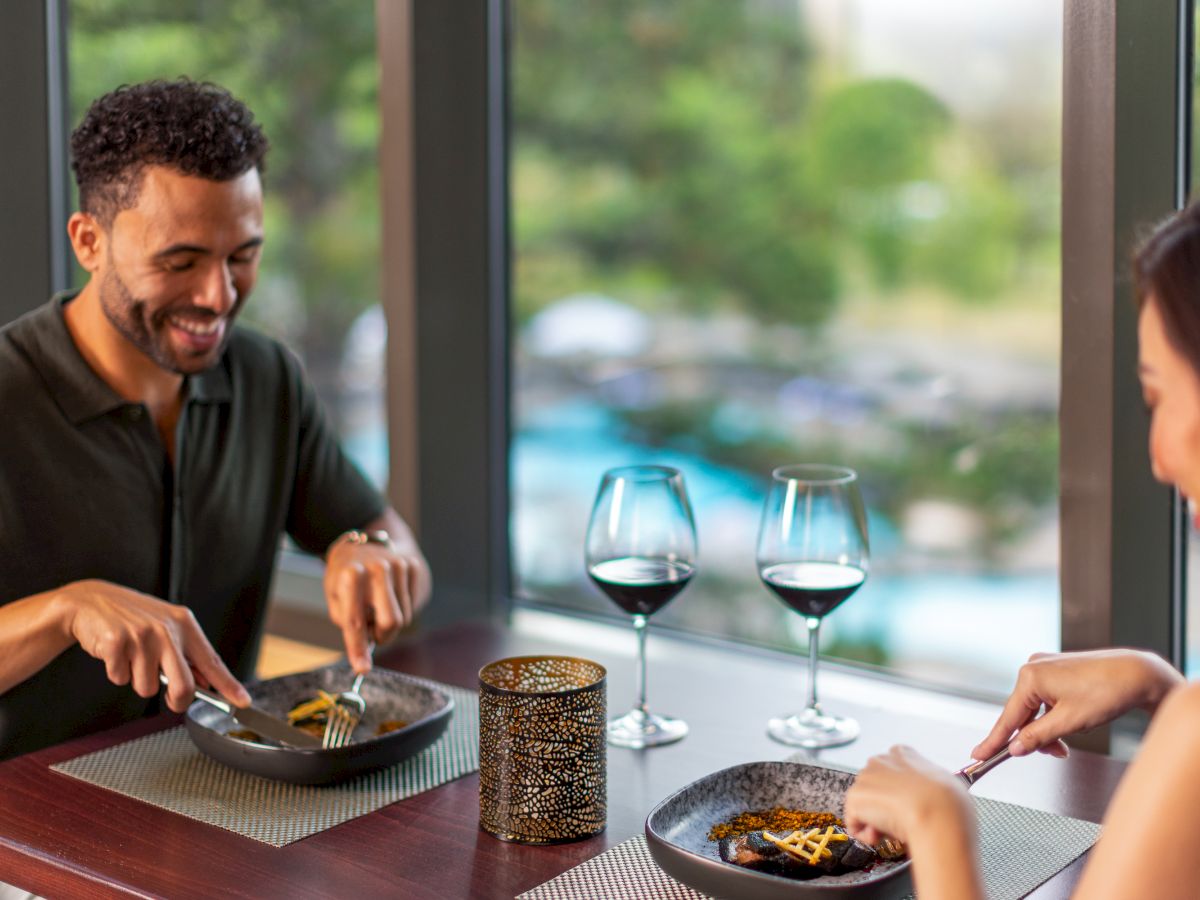 A couple is dining, enjoying pasta with red wine near a window. A candle is on the table, providing a cozy atmosphere.