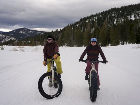 Two people are riding fat bikes on a snowy trail, surrounded by trees and mountains under a cloudy sky.