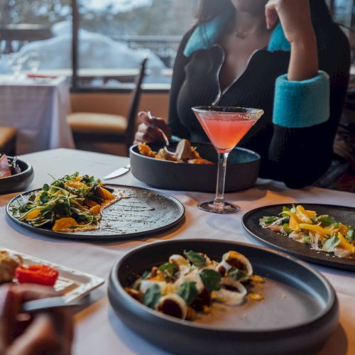 A dining table with various dishes and a cocktail; a person seated in a cozy restaurant setting.