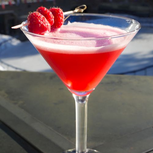 A vibrant red cocktail in a martini glass garnished with raspberries on a skewer, set against an outdoor backdrop.