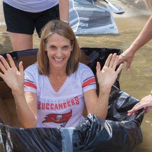 A woman smiles and waves while riding in a black tube through shallow muddy water at a camp, with friends holding the raft.