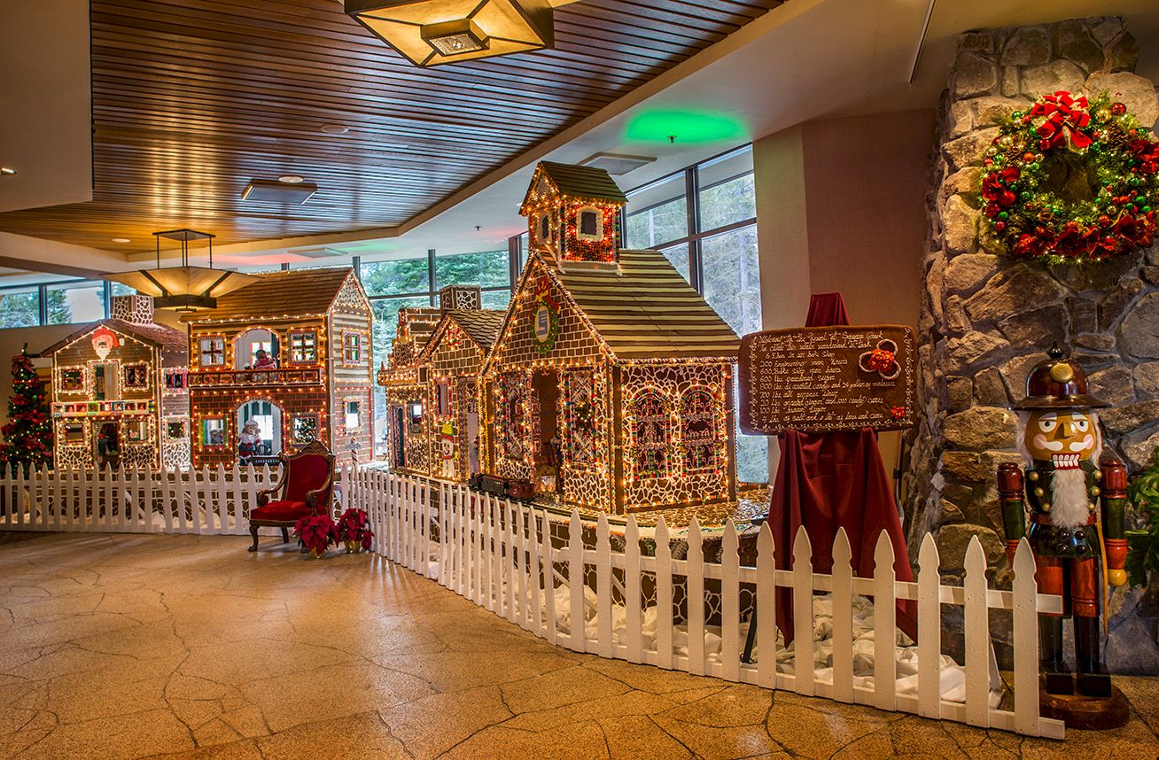 Indoor holiday display of glowing gingerbread-style houses, a nutcracker, wreaths, and festive lights along a fence.