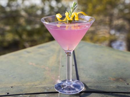 A pink cocktail in a martini glass, garnished with lemon twist and rosemary, set against a blurred outdoor background.