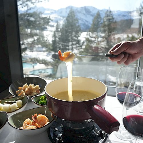 A fondue set with cheese, surrounded by bowls of food and glasses of wine, overlooks a snowy mountain view from a window.