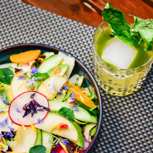 A colorful salad with sliced veggies and edible flowers, plus a glass of bright green mint drink over ice.