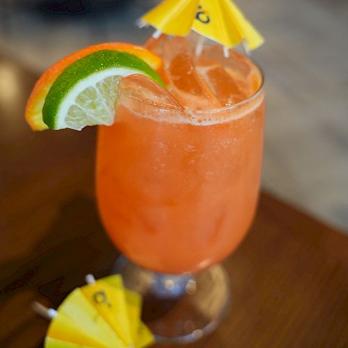 A tropical orange cocktail in a glass with ice, garnished with orange and lime slices and a yellow umbrella on top and side.