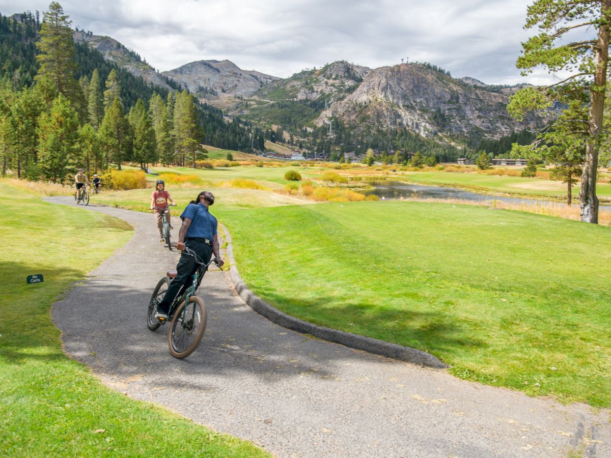 Cyclists ride along a lakeside path through a grassy valley with towering mountains and trees under cloudy skies.