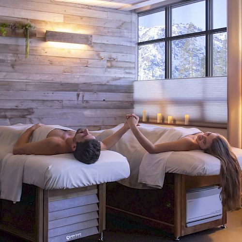 A serene spa room with two people relaxing on massage tables, holding hands, surrounded by candles and a window view of snowy mountains.