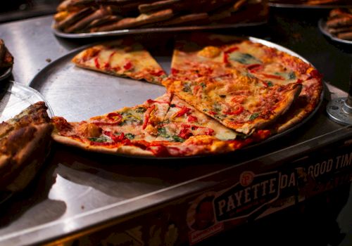 A pizza with several slices is on a metal tray, topped with red peppers and herbs, placed on a counter with partial text visible at the front.