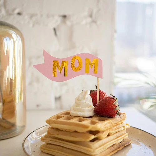 A stack of waffles topped with whipped cream and strawberries, a pink "MOM" sign on top, set against a light background.