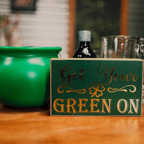 A green pot, a bottle, glass mugs, and a sign saying "Get Your Green On" are on a wooden surface, suggesting a festive theme.