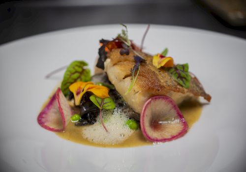 A plated dish featuring seared fish, colorful vegetables, sauce, microgreens, and edible flowers on a white plate.