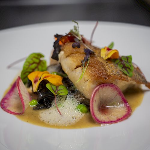 A plated dish featuring seared fish, colorful vegetables, sauce, microgreens, and edible flowers on a white plate.