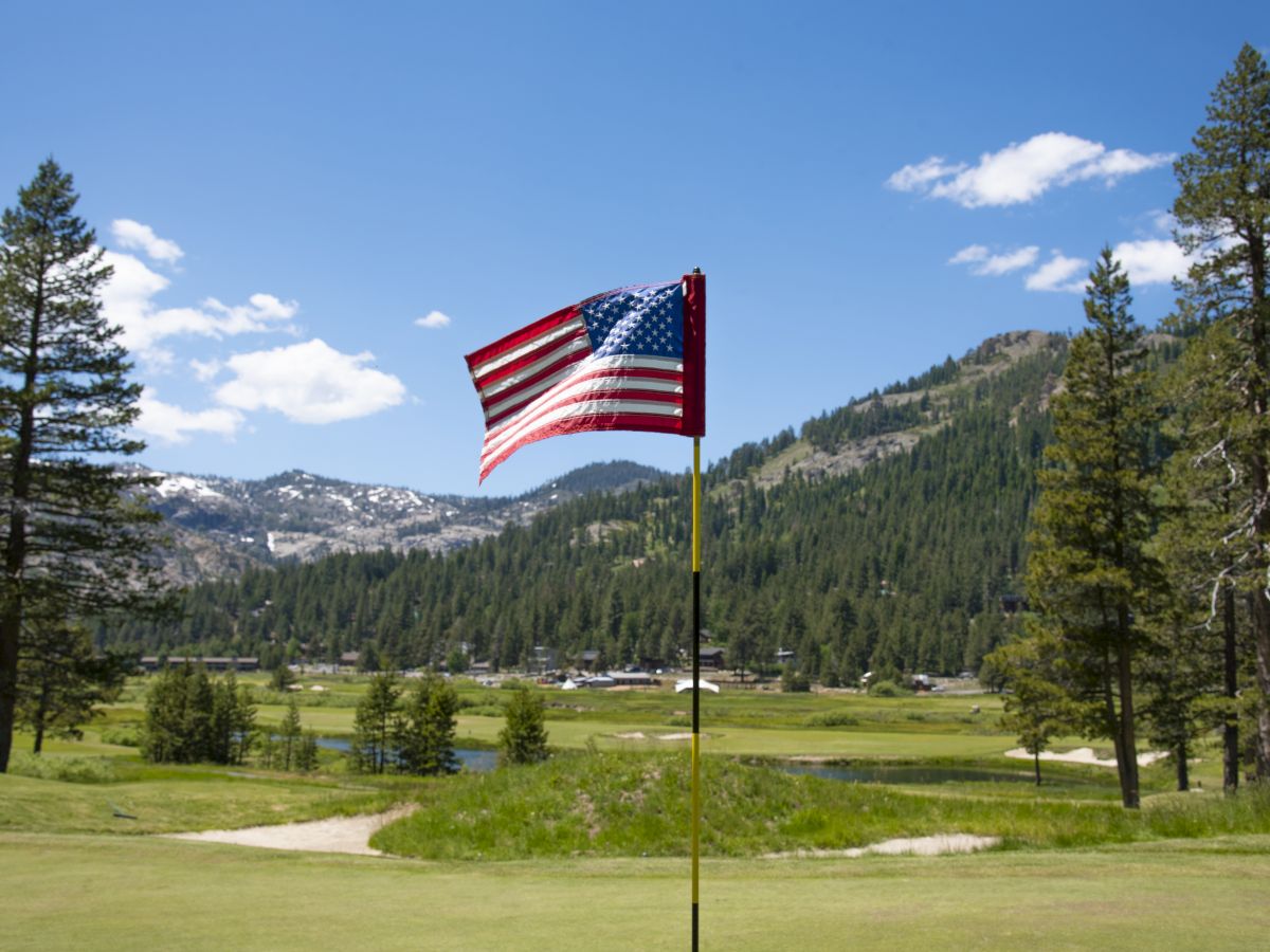 A golf course with an American flag on the hole, surrounded by trees and mountains under a bright blue sky with clouds.