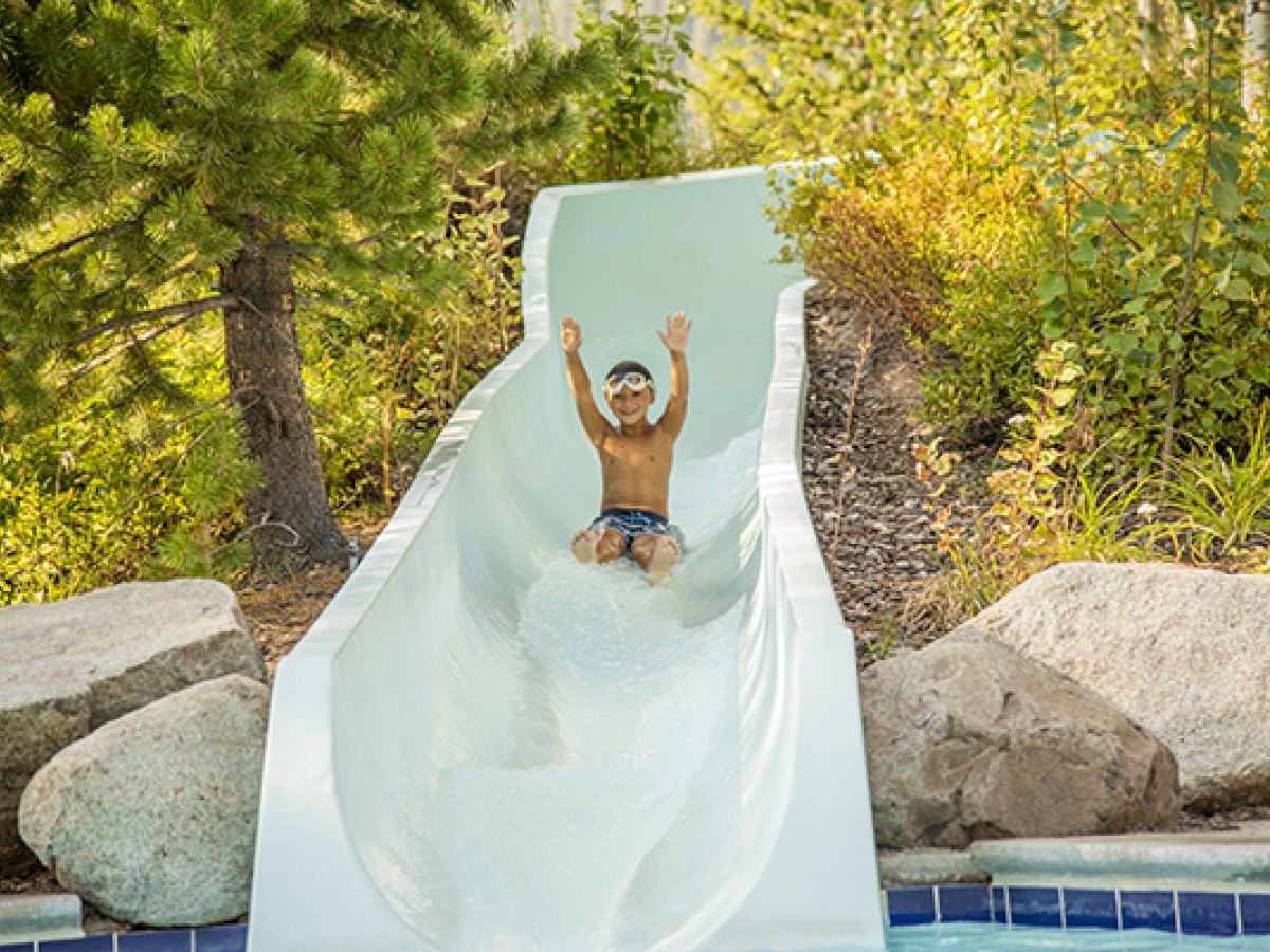 A child wearing goggles slides down a water slide with arms raised, surrounded by trees and rocks, with a pool at the bottom.