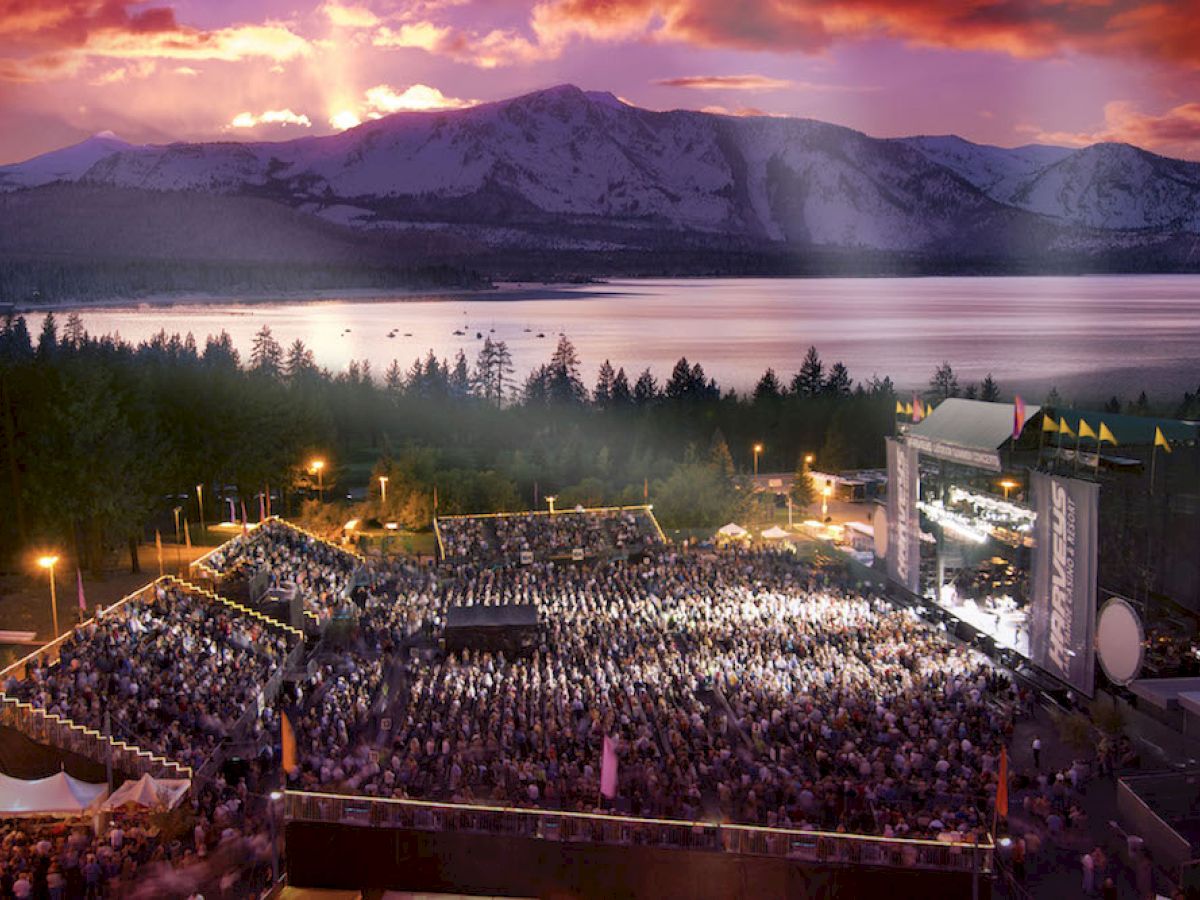 Outdoor concert with large crowd, stage, and dramatic sunset view over mountains and a lake.
