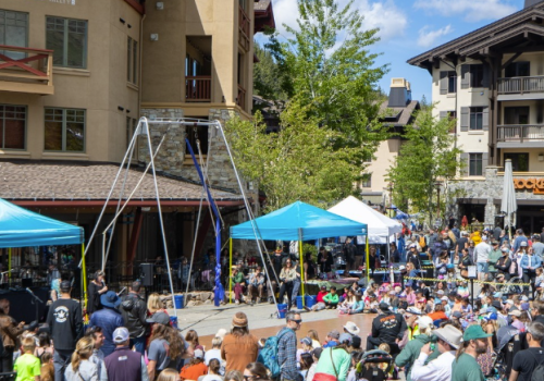 Outdoor event with many people, blue tents, and a climbing structure in a plaza surrounded by buildings and trees under a blue sky.