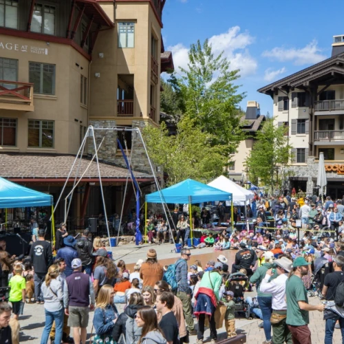Outdoor event with many people, blue tents, and a climbing structure in a plaza surrounded by buildings and trees under a blue sky.