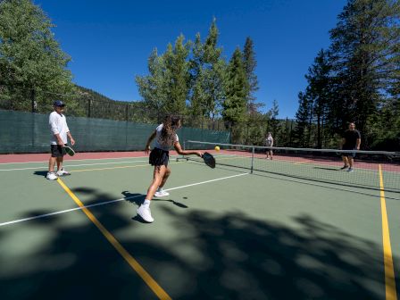 People are playing pickleball on an outdoor court surrounded by trees under a clear blue sky.