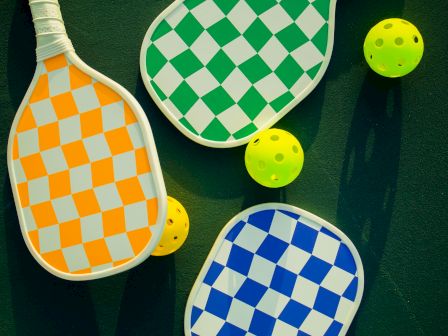 The image shows three paddles with checkered patterns in orange, green, and blue, alongside three yellow pickleballs.