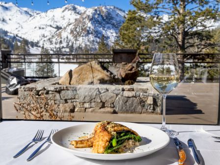 A meal with salmon and veggies on a table, accompanied by white wine, with a snowy mountain view in the background.