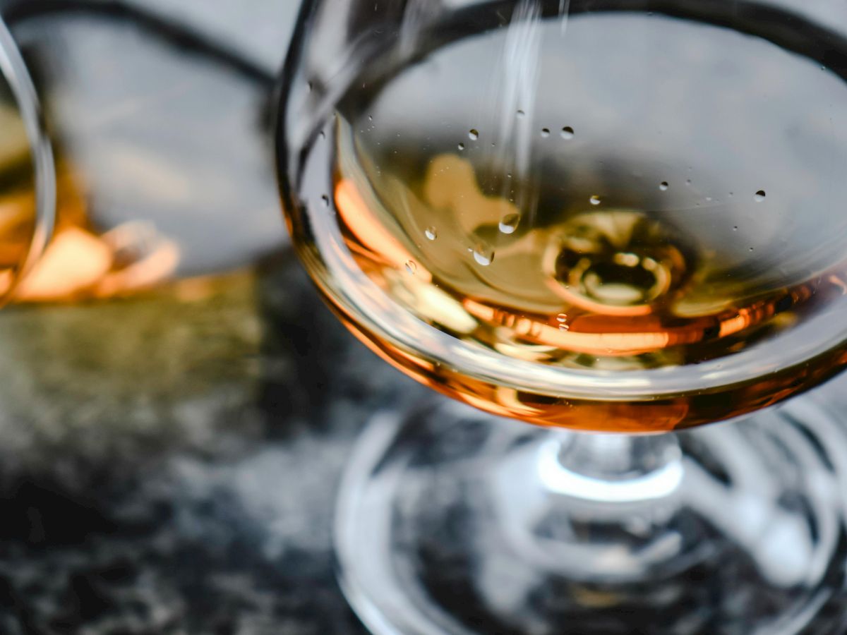 A close-up of a glass of amber liquid, likely whiskey or brandy, on a textured surface, with another glass blurred in the background.