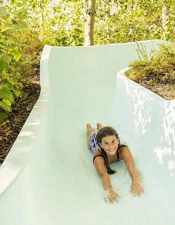 A child is sliding down a slide in a natural setting surrounded by lush green trees, smiling and enjoying the ride through nature.