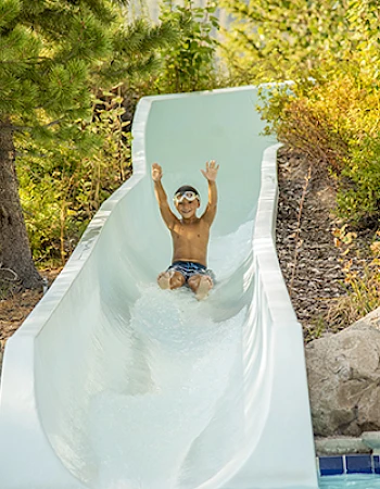 A child wearing goggles slides down a water slide with arms raised, surrounded by trees and rocks, heading toward a pool.