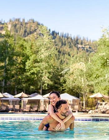 A couple enjoys fun in a pool, surrounded by trees and mountains. Umbrellas and tents are in the background, creating a relaxing setting.