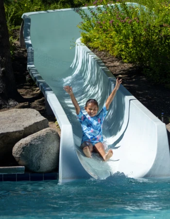 A child joyfully slides down a water slide into a pool, surrounded by greenery and rocks, arms raised in excitement.