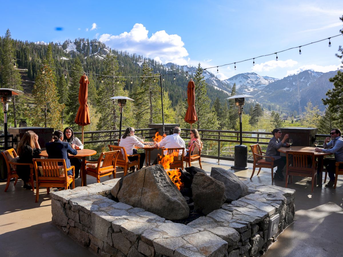 People are sitting around a fire pit on a sunny patio with mountains in the background.