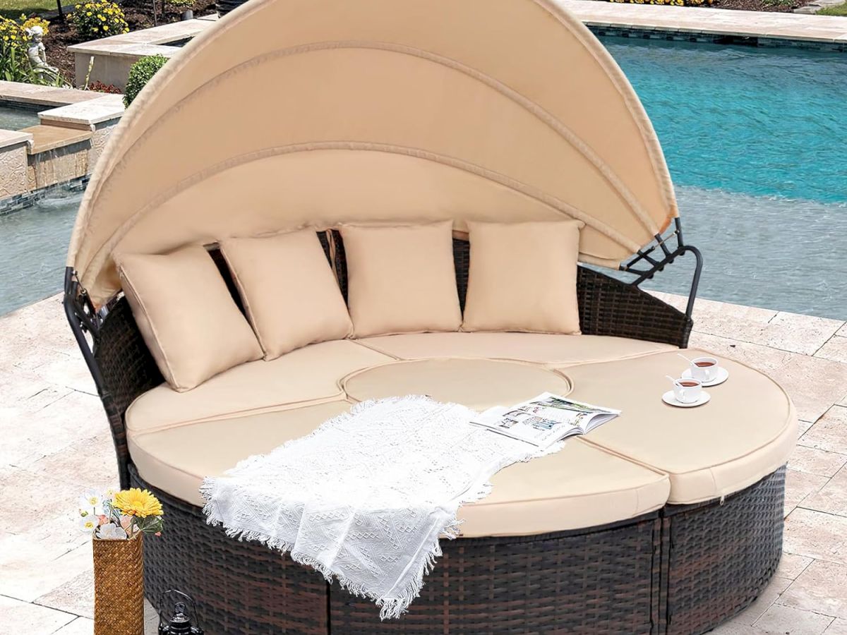 A round outdoor daybed with a canopy and beige cushions is by a pool, featuring pillows, a magazine, and drinks on the side.